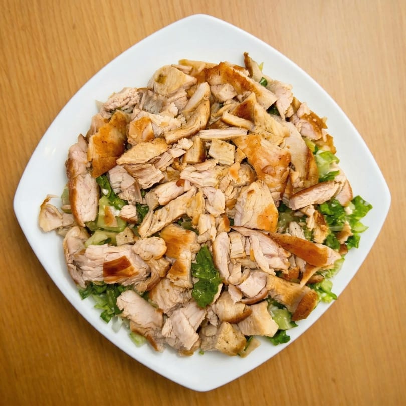 Overhead view of a white square plate filled with chopped chicken and green salad on a wooden table.