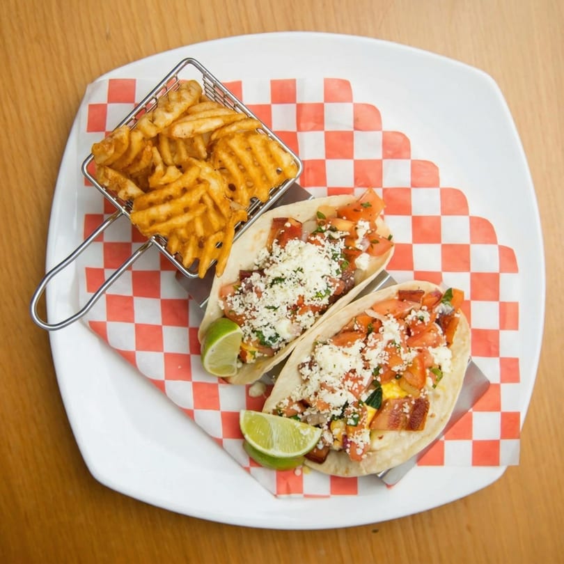 Two tacos with cheese, tomatoes, and lime wedges, served with waffle fries in a metal basket.