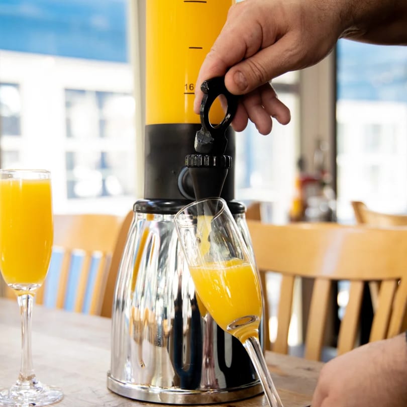 A hand pouring orange juice from a large dispenser into a champagne flute at Gramercy Kitchen.