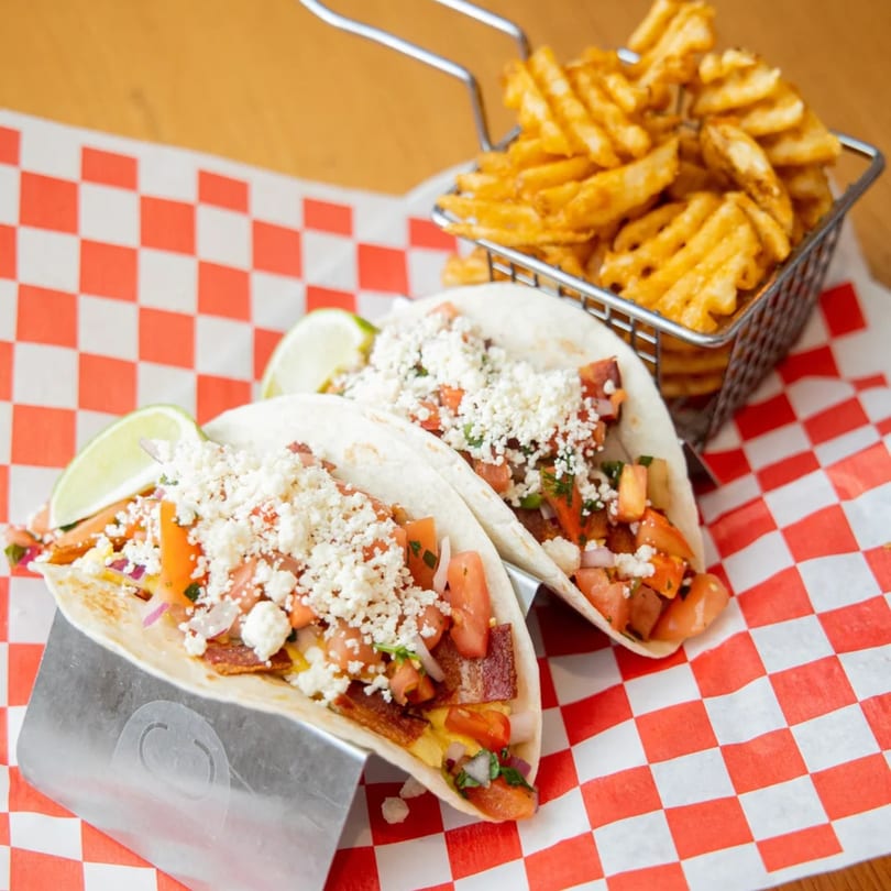 Two breakfast tacos with scrambled eggs, bacon, pico de gallo, and feta, served with waffle fries.